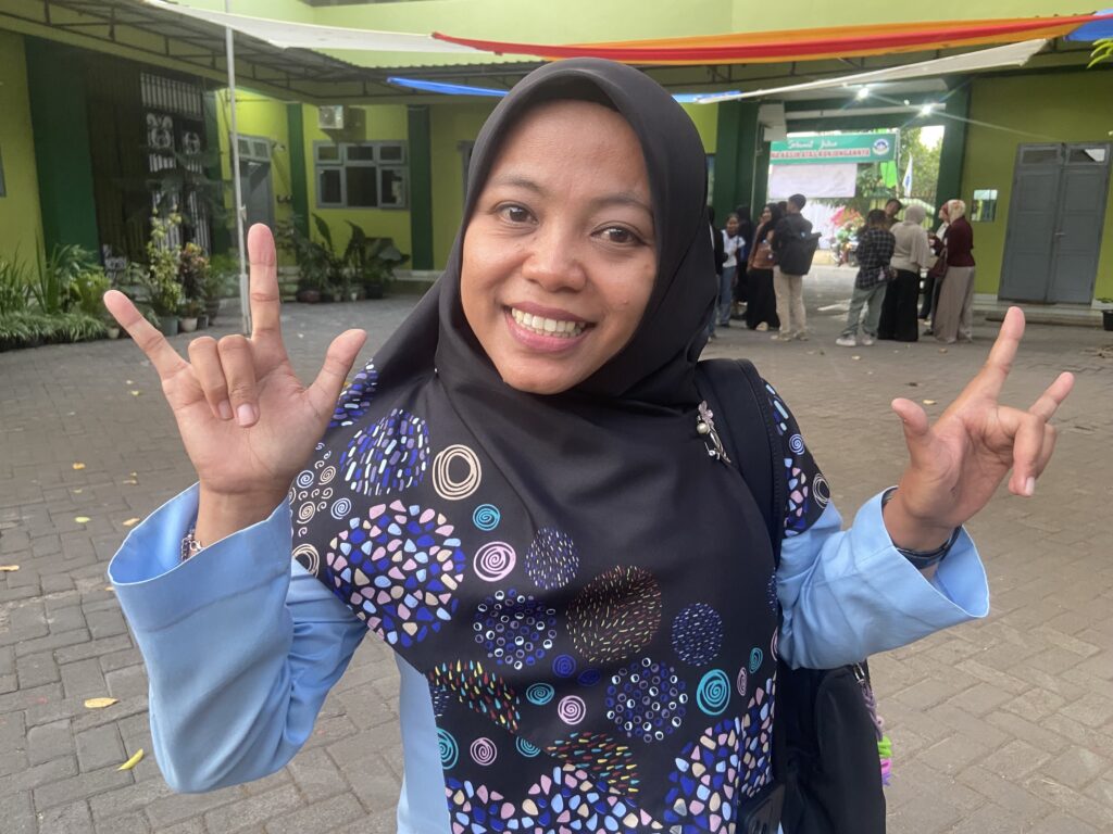 Smiling woman in a black hijab and blue patterned dress takes a selfie, hands raised in rock-on gestures, outdoors with people in the background.