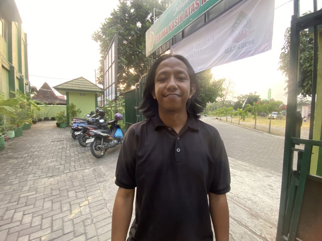 Person in a black shirt standing outdoors on a brick-paved path, with a green fence and parked motorcycles in the background.
