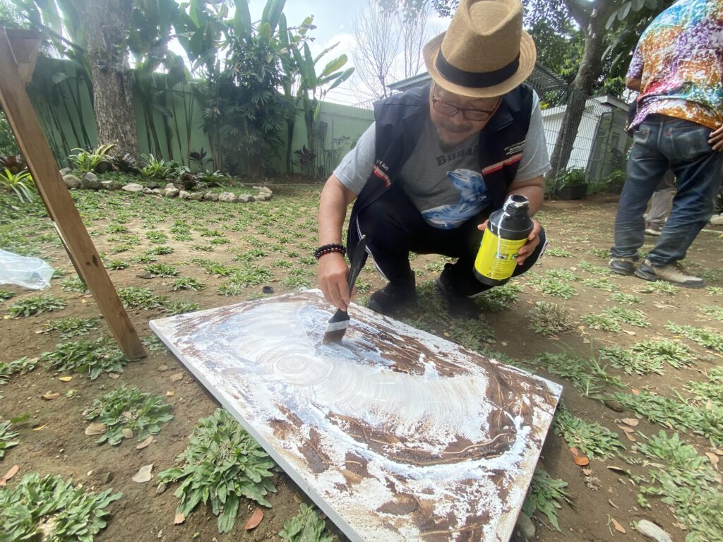 IMG_1083 - Mabur.co Man in a straw hat and glasses kneels outdoors, painting a large board with brown paint, holding a yellow paint bottle.