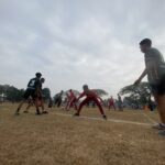Rugby players contest the ball on a dusty field, wearing green and red uniforms, with a coach or spectator at the right edge.