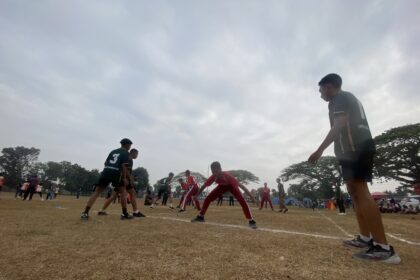 Rugby players contest the ball on a dusty field, wearing green and red uniforms, with a coach or spectator at the right edge.