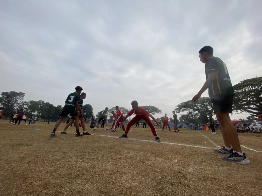 Rugby players contest the ball on a dusty field, wearing green and red uniforms, with a coach or spectator at the right edge.