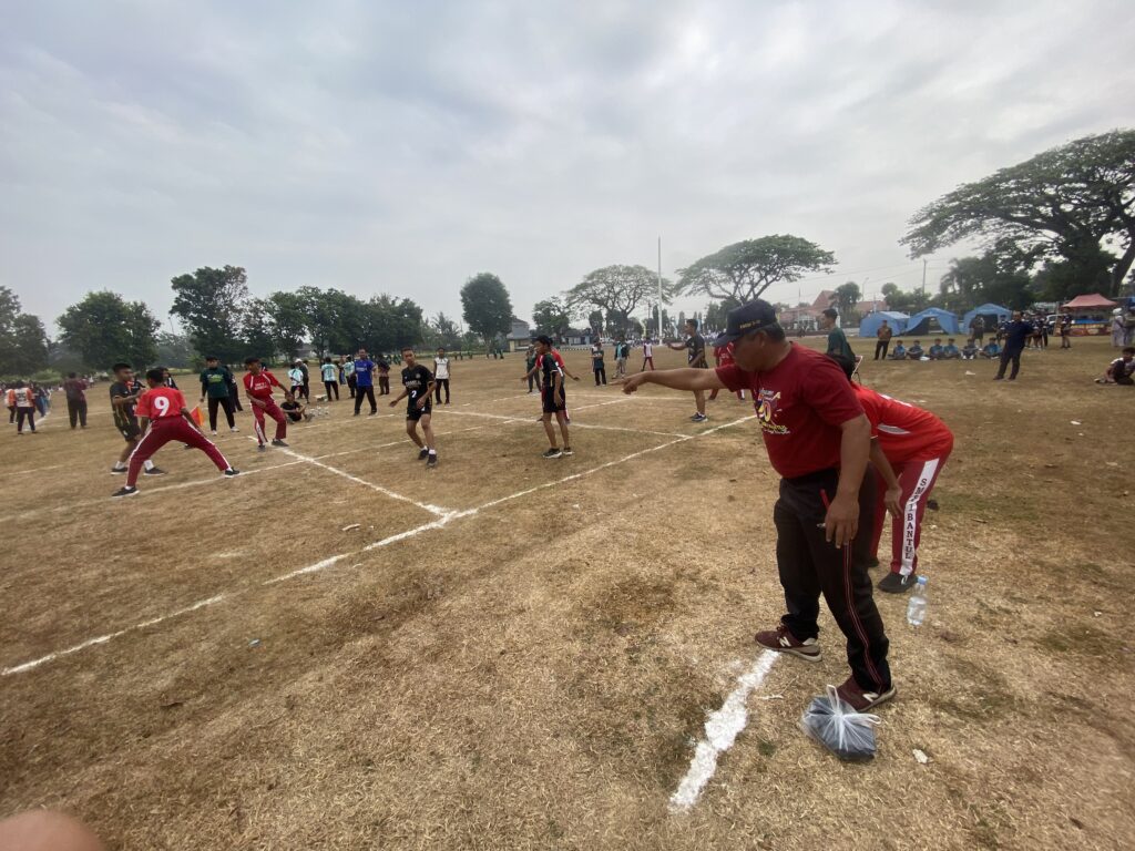 IMG_1113 - Mabur.co Coach in red gestures toward the field as young players in colorful jerseys run and practice on a dirt sports field, with tents and trees in the background.