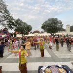 Large crowd in colorful traditional clothing at an outdoor festival, with trees and a decorative stage to the side.