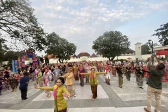 Large crowd in colorful traditional clothing at an outdoor festival, with trees and a decorative stage to the side.