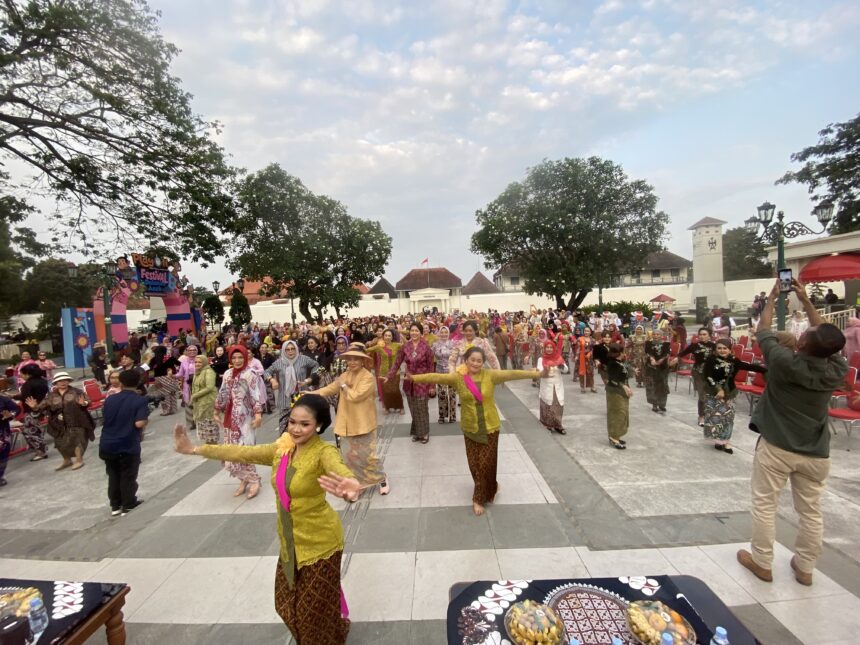 Large crowd in colorful traditional clothing at an outdoor festival, with trees and a decorative stage to the side.