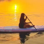 Person paddling a white stand-up paddleboard on calm water at sunset, orange sky with sun reflection on the surface.