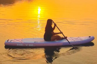 Person paddling a white stand-up paddleboard on calm water at sunset, orange sky with sun reflection on the surface.
