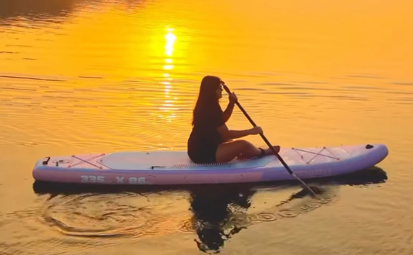 Person paddling a white stand-up paddleboard on calm water at sunset, orange sky with sun reflection on the surface.