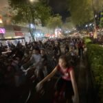 Nighttime street scene with a dense crowd of cyclists and pedestrians, storefronts lit up along a tree-lined avenue.
