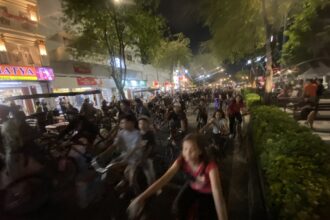 Nighttime street scene with a dense crowd of cyclists and pedestrians, storefronts lit up along a tree-lined avenue.