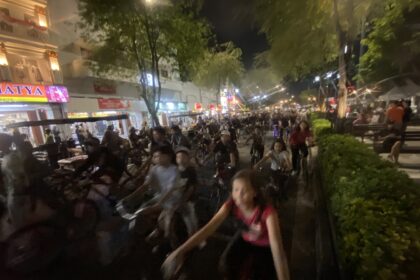 Nighttime street scene with a dense crowd of cyclists and pedestrians, storefronts lit up along a tree-lined avenue.