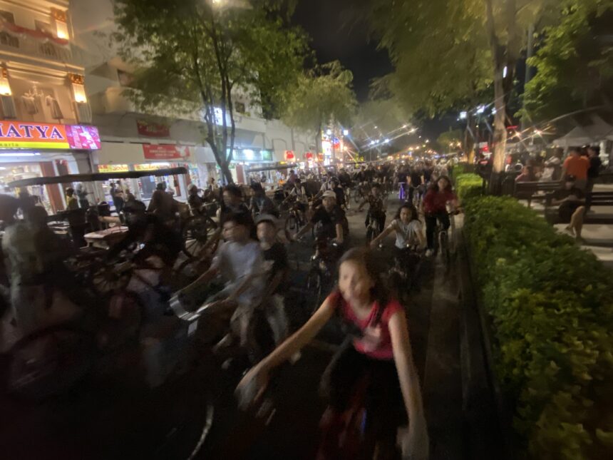 Nighttime street scene with a dense crowd of cyclists and pedestrians, storefronts lit up along a tree-lined avenue.