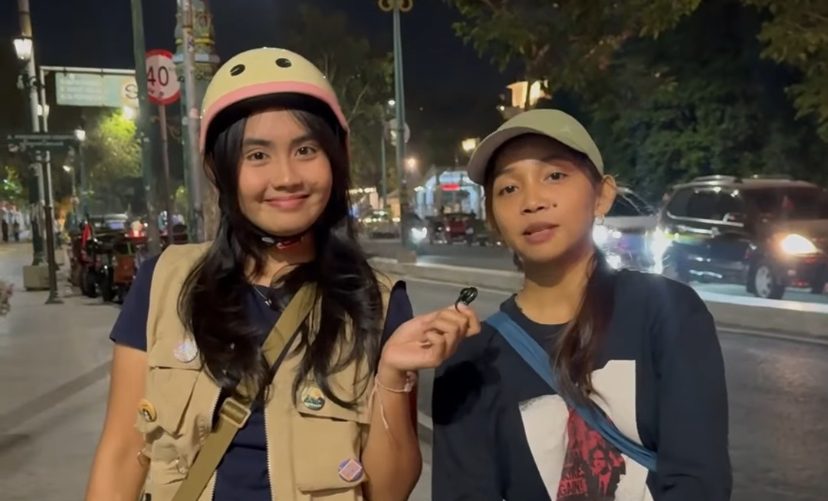 IMG_1281 - Mabur.co Two young women stand on a night city sidewalk; one wears a beige vest and a pink helmet, the other a cap, both smiling, with the first holding a small object.