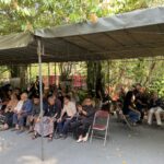 Group of people seated on folding chairs under a large outdoor canopy with trees and plants around.