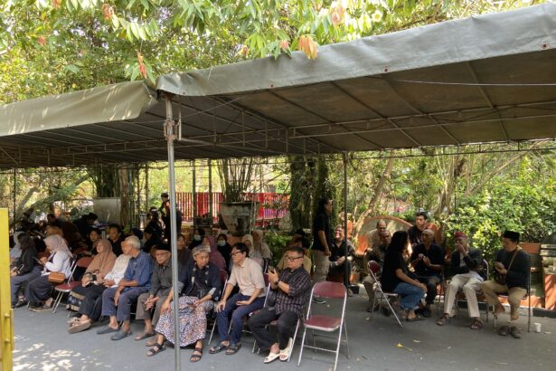 Group of people seated on folding chairs under a large outdoor canopy with trees and plants around.