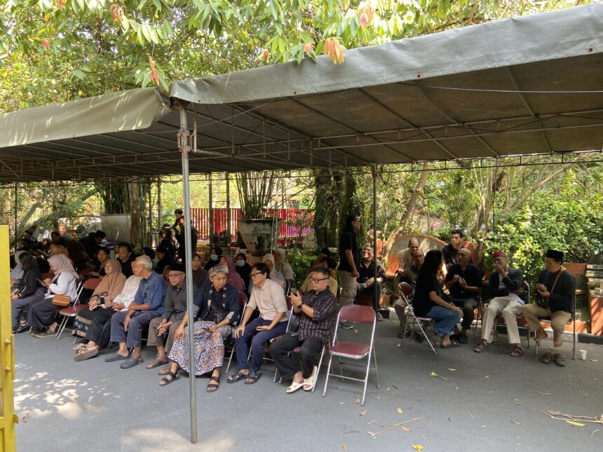Group of people seated on folding chairs under a large outdoor canopy with trees and plants around.
