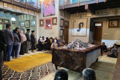 A group of people in a decorated room standing and seated for a funeral ritual; a body lies under a patterned cloth on a raised table, with attendees in modest clothing and head coverings nearby