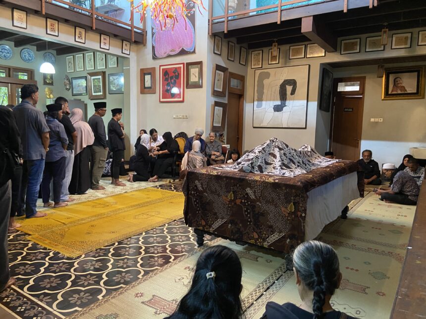 A group of people in a decorated room standing and seated for a funeral ritual; a body lies under a patterned cloth on a raised table, with attendees in modest clothing and head coverings nearby