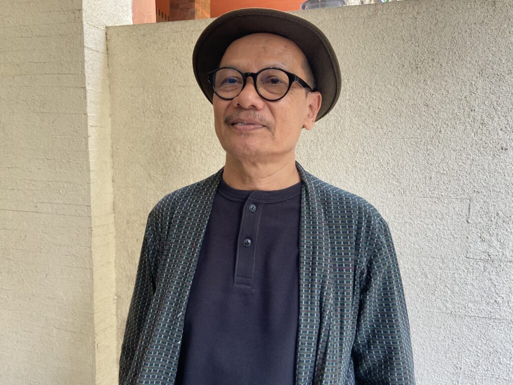 IMG_1330 - Mabur.co Portrait of a man in a dark shirt and checkered blazer wearing round glasses and a brown hat, standing against a beige wall.