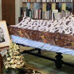 Funeral display with a framed portrait of an elderly man, floral arrangements, and a casket covered by patterned fabric in a room filled with bookshelves