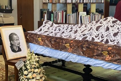 Funeral display with a framed portrait of an elderly man, floral arrangements, and a casket covered by patterned fabric in a room filled with bookshelves