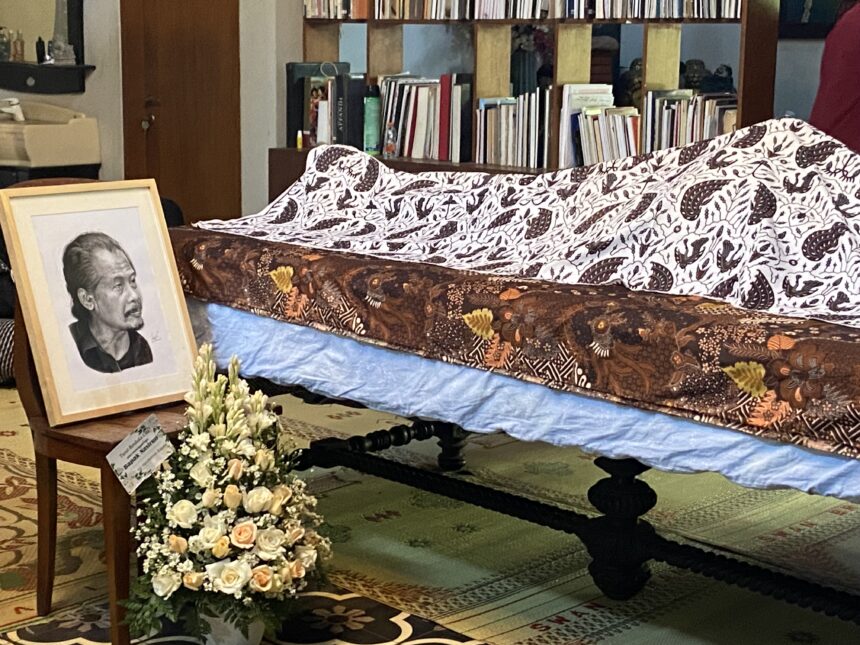 Funeral display with a framed portrait of an elderly man, floral arrangements, and a casket covered by patterned fabric in a room filled with bookshelves