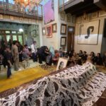 Crowd gathered in a gallery-like room with framed art on the walls and long tables covered in a black-and-white patterned cloth displaying photographs or portraits.