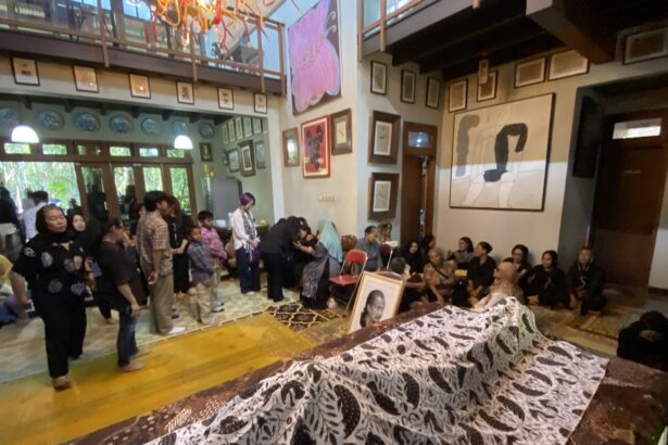 Crowd gathered in a gallery-like room with framed art on the walls and long tables covered in a black-and-white patterned cloth displaying photographs or portraits.