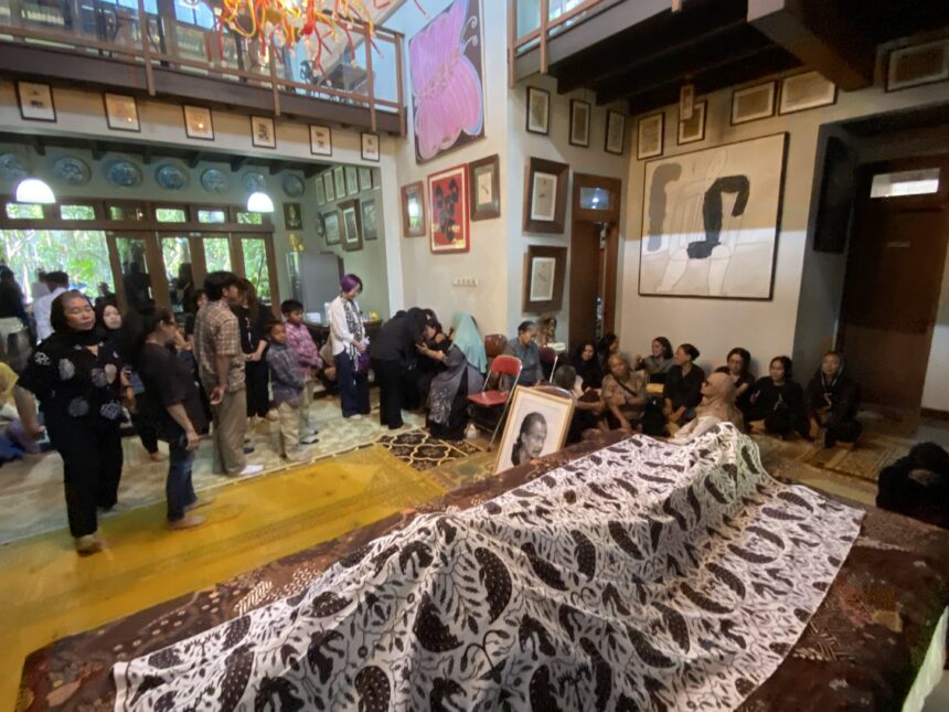 Crowd gathered in a gallery-like room with framed art on the walls and long tables covered in a black-and-white patterned cloth displaying photographs or portraits.