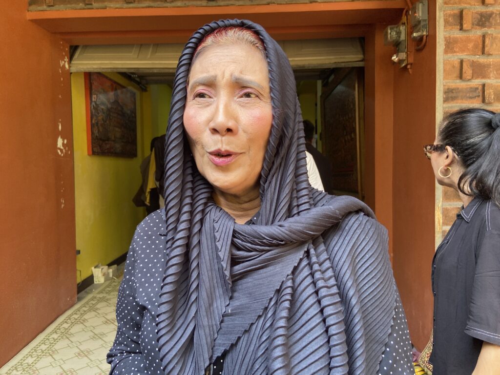 IMG_1351 - Mabur.co Close-up of an older woman wearing a dark, ribbed scarf outdoors near a brick doorway, with another person partially visible on the right.