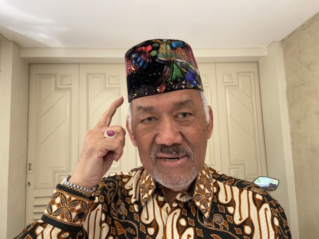 IMG_1370 - Mabur.co Older man with a multicolored, paint-splattered fez hat, touching his temple indoors.