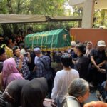 Green coffin draped with tassels being carried through a crowded outdoor procession by mourners holding phones and people watching.