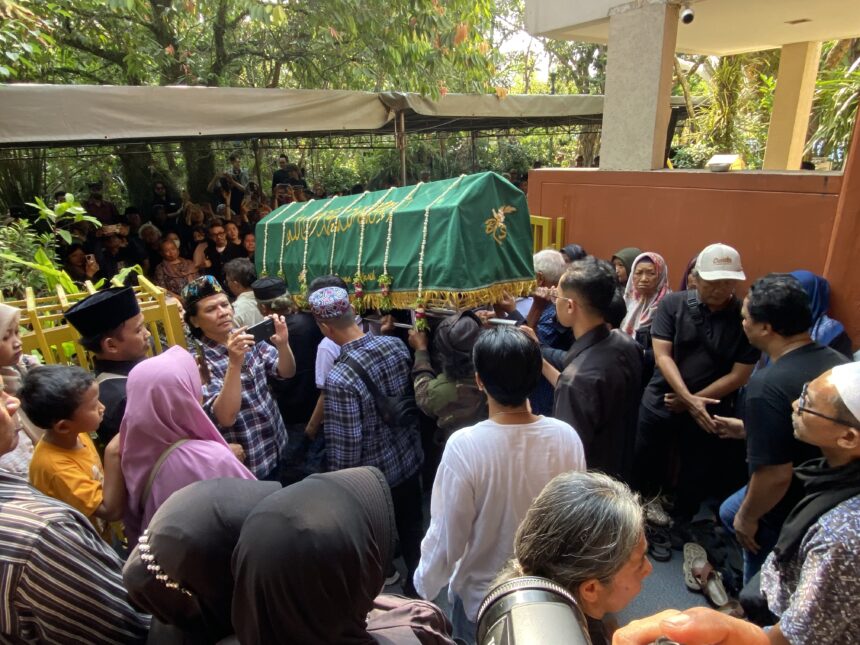 Green coffin draped with tassels being carried through a crowded outdoor procession by mourners holding phones and people watching.