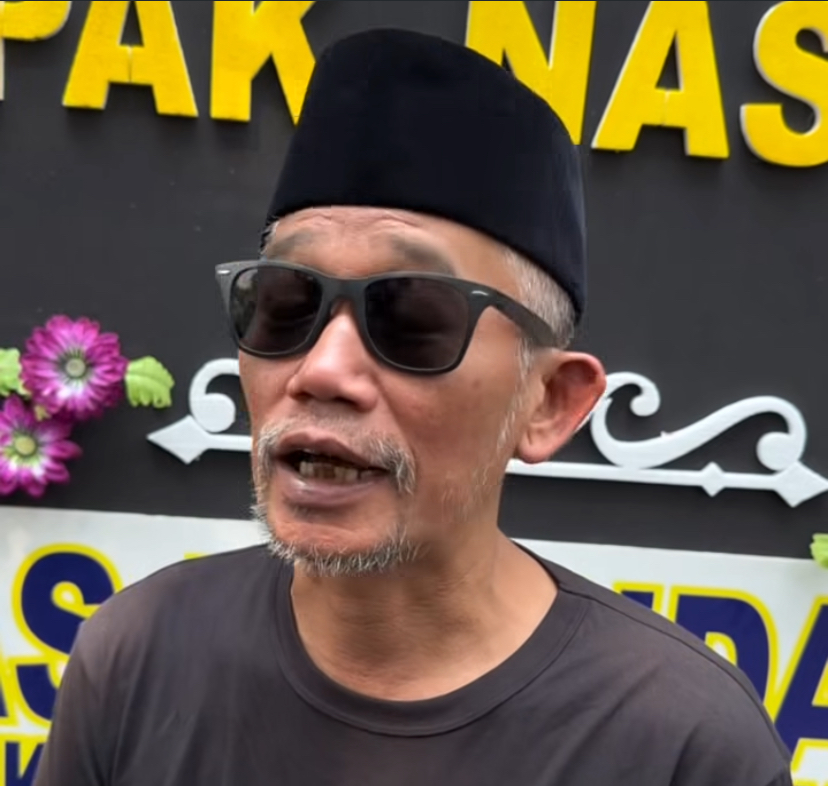 IMG_1384 - Mabur.co Man wearing a black cap and sunglasses speaks in front of a sign with yellow letters in the background.