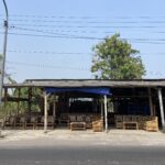 Open-air furniture workshop with many wooden bamboo chairs under a corrugated roof, power lines overhead.