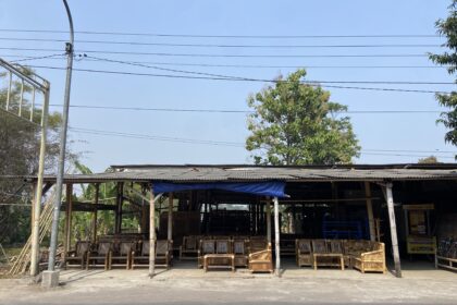 Open-air furniture workshop with many wooden bamboo chairs under a corrugated roof, power lines overhead.