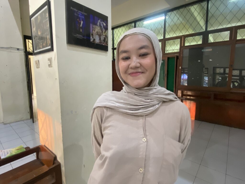 Woman wearing a beige hijab and top smiles in a indoor hallway near glass doors and posters on the wall