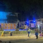 Nighttime outdoor stage performance with dancers in traditional blue-green costumes under blue lighting, ropes and banners in the foreground.