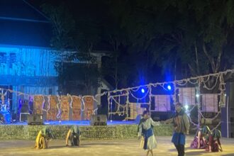 Nighttime outdoor stage performance with dancers in traditional blue-green costumes under blue lighting, ropes and banners in the foreground.