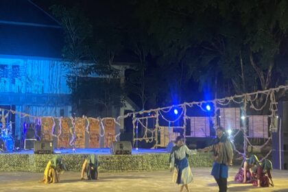 Nighttime outdoor stage performance with dancers in traditional blue-green costumes under blue lighting, ropes and banners in the foreground.