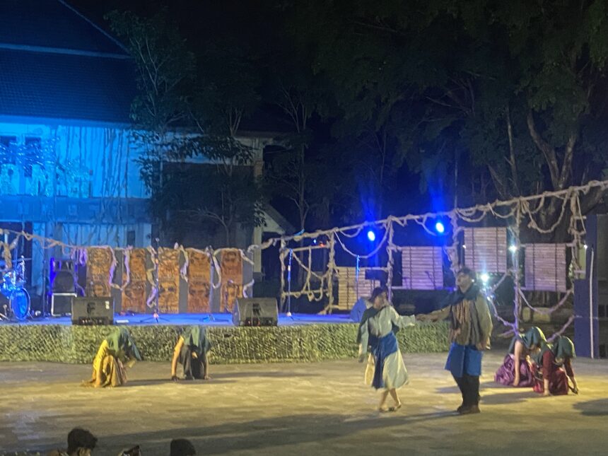 Nighttime outdoor stage performance with dancers in traditional blue-green costumes under blue lighting, ropes and banners in the foreground.