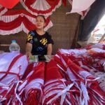 Woman sitting under a red-and-white fabric canopy, surrounded by piles of red and white bundles at a market stall.