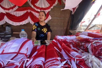 Woman sitting under a red-and-white fabric canopy, surrounded by piles of red and white bundles at a market stall.