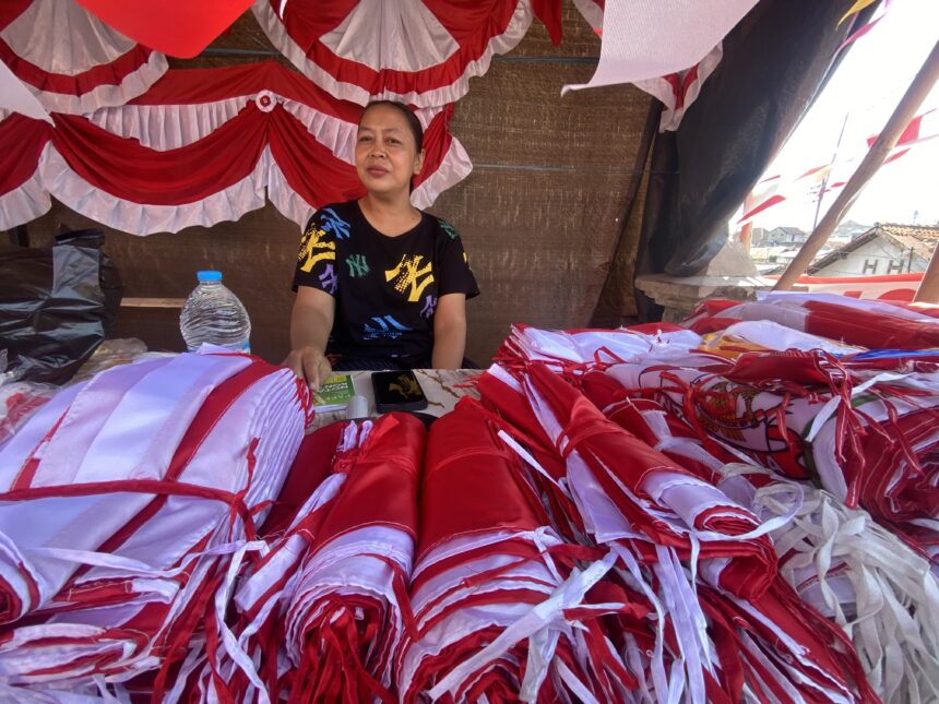 Woman sitting under a red-and-white fabric canopy, surrounded by piles of red and white bundles at a market stall.