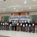 Group of men and women in batik attire on a stage, each holding a certificate during a cultural village awards ceremony