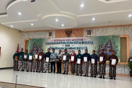 Group of men and women in batik attire on a stage, each holding a certificate during a cultural village awards ceremony