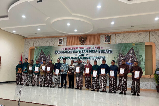Group of men and women in batik attire on a stage, each holding a certificate during a cultural village awards ceremony