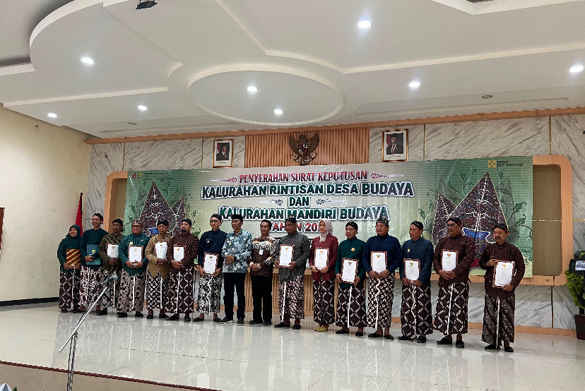 Group of men and women in batik attire on a stage, each holding a certificate during a cultural village awards ceremony