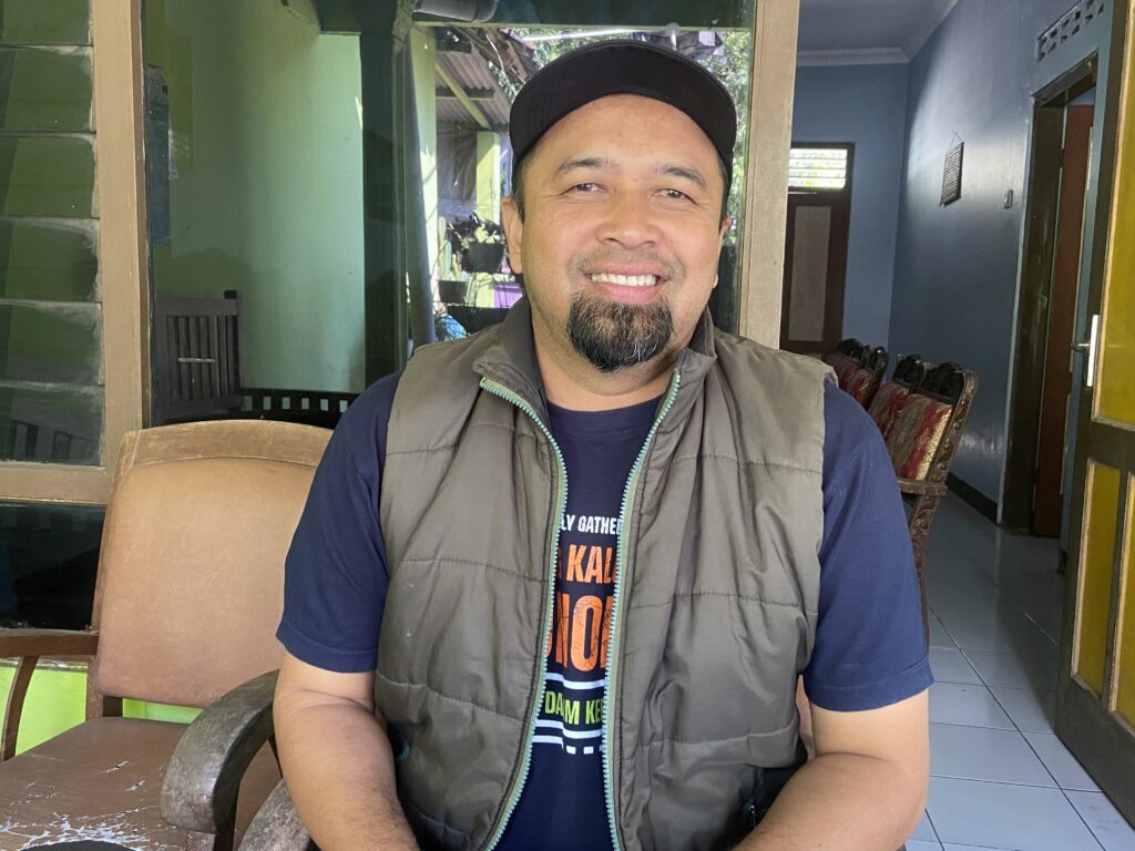 Smiling man wearing a black cap and olive vest seated indoors, cafe seating and green walls visible behind him.